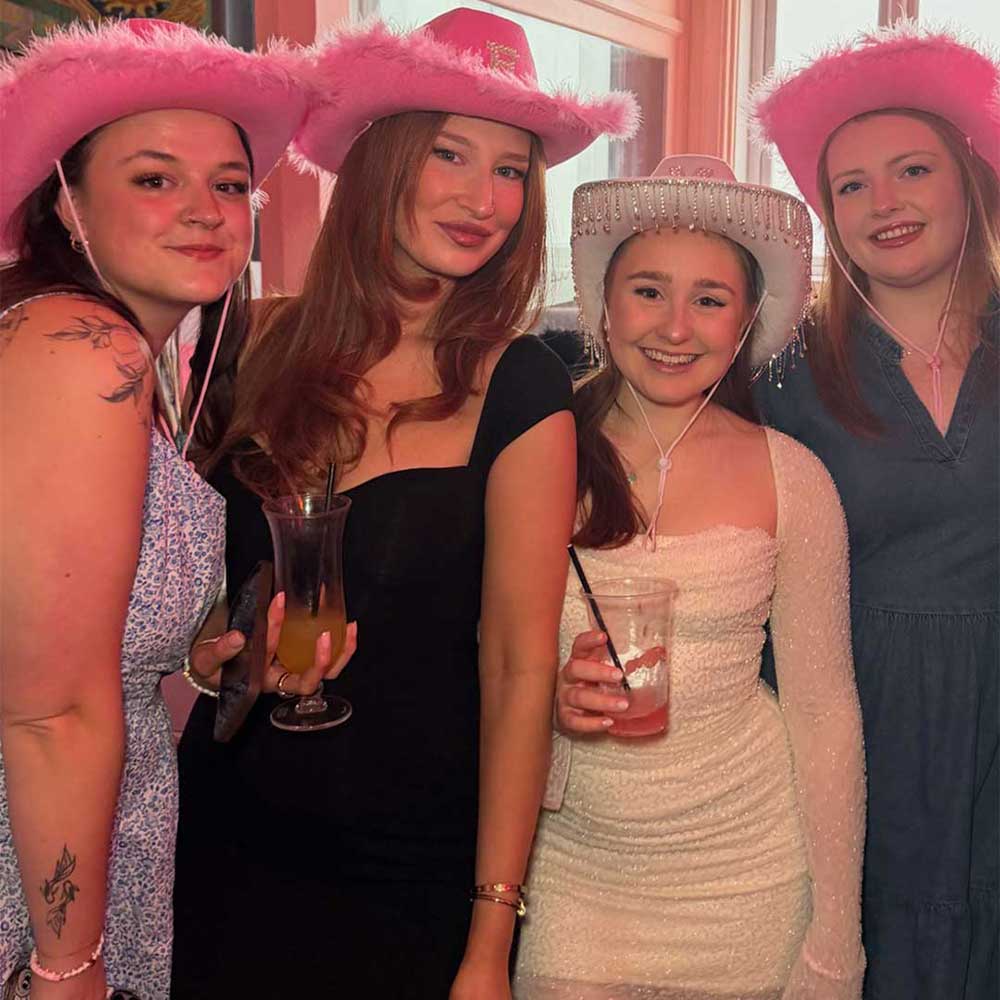 Four women wearing pink hats with feather embellishments, holding drinks, and posing together at a themed bottomless brunch event in Greenwich for Lovebrunch